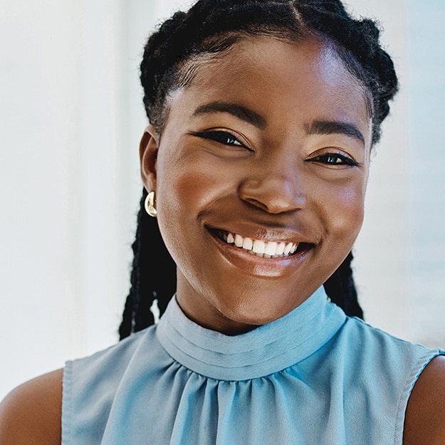 Smiling young woman with dark braided hair and glowing skin wearing a light blue high-neck sleeveless top against a bright background.
