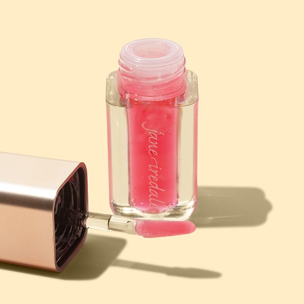 jane iredale lip oil in a sheer pink shade with its cushion applicator and rose gold cap on a cream background.