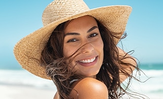 A smiling woman wearing a wide-brim sun hat poses on a sunny beach.