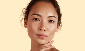 A woman with radiant, glowing skin poses against a soft yellow background.