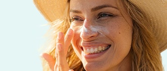 Smiling young woman wearing a sunhat and applying sunscreen to her face.