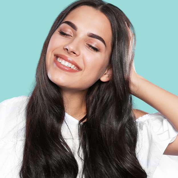 Smiling woman with long dark hair and eyes closed, hands lifted to her face, against a teal background.