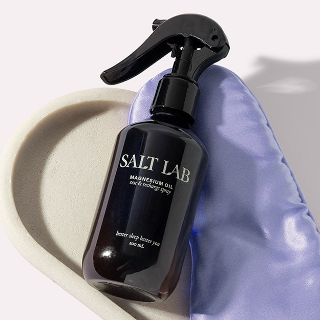 Salt Lab Spray bottle resting on a satin pillow beside a cream stone dish.