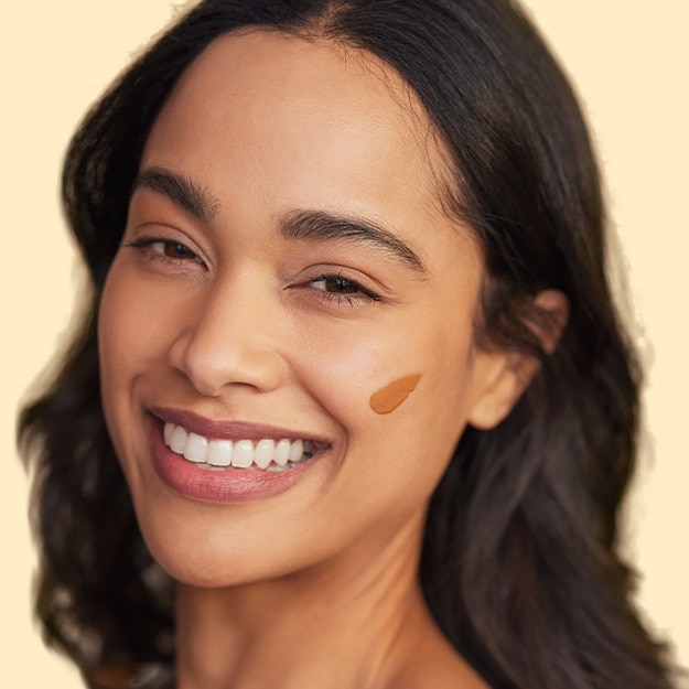 Smiling woman with curly light-brown hair applying a pink powder brush to her cheek.