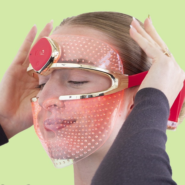 Model wearing red LED light face mask device against a light green background.