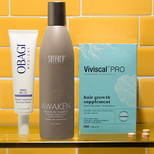 Obagi Nu-Cil scalp serum tube, Surface Awaken Therapeutic Shampoo bottle and a Viviscal PRO hair growth supplement box sit on a glass shelf.