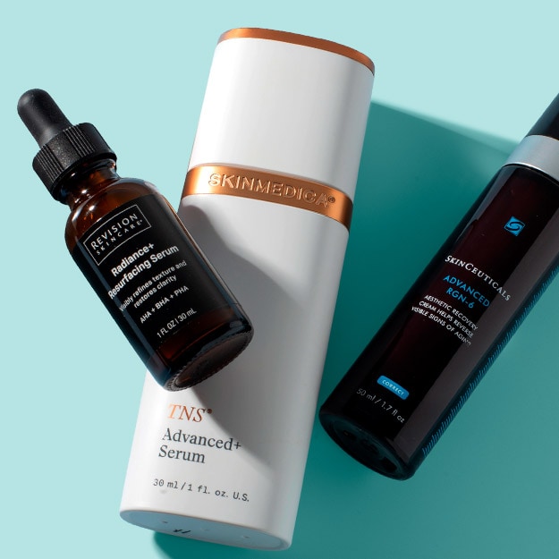 Three anti-aging serums from Revision Skincare, SkinMedica and SkinCeuticals displayed on a light blue surface