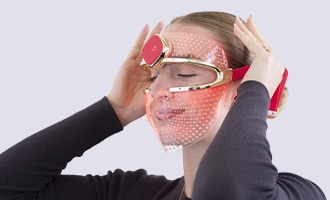 Person wearing red LED light therapy mask with hands positioned on either side of face