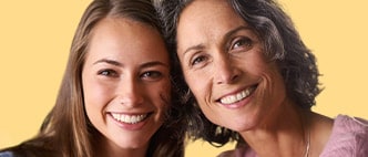 Two smiling women (mother and daughter) posing together against a warm yellow background.